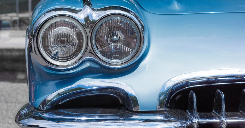 A close-up of an older Corvette car in a gray-blue color. The two front headlights are off and shiny.