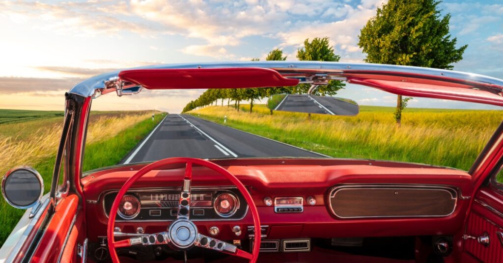 View from red vintage convertible driving down country road, trees and fields under blue sky with scattered clouds.