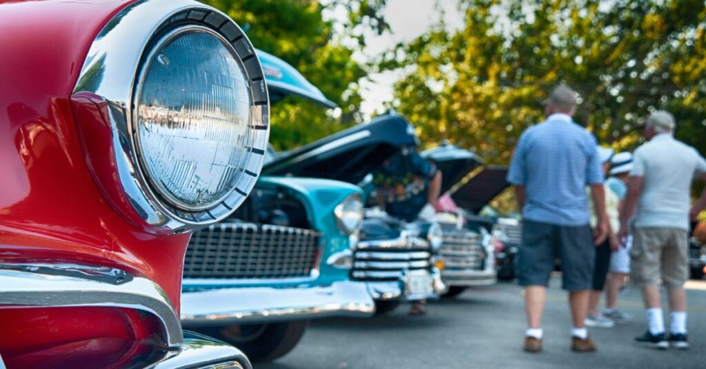 Close-up of a red classic car headlight with other vintage cars and visitors at an outdoor car show.