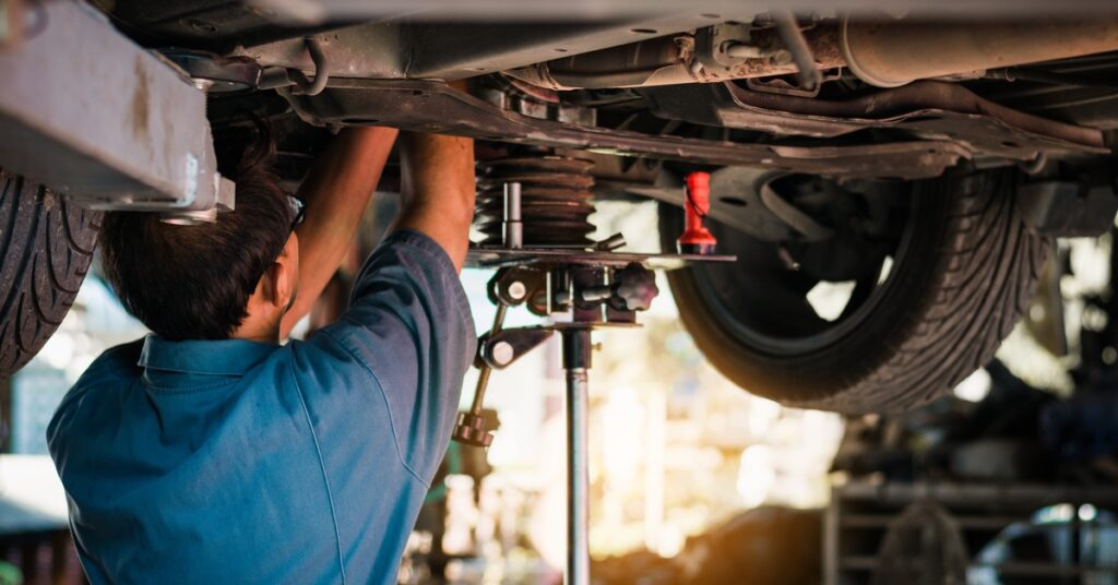 Mechanic works under raised car on lift, reaching toward suspension and wheel components in auto repair shop.