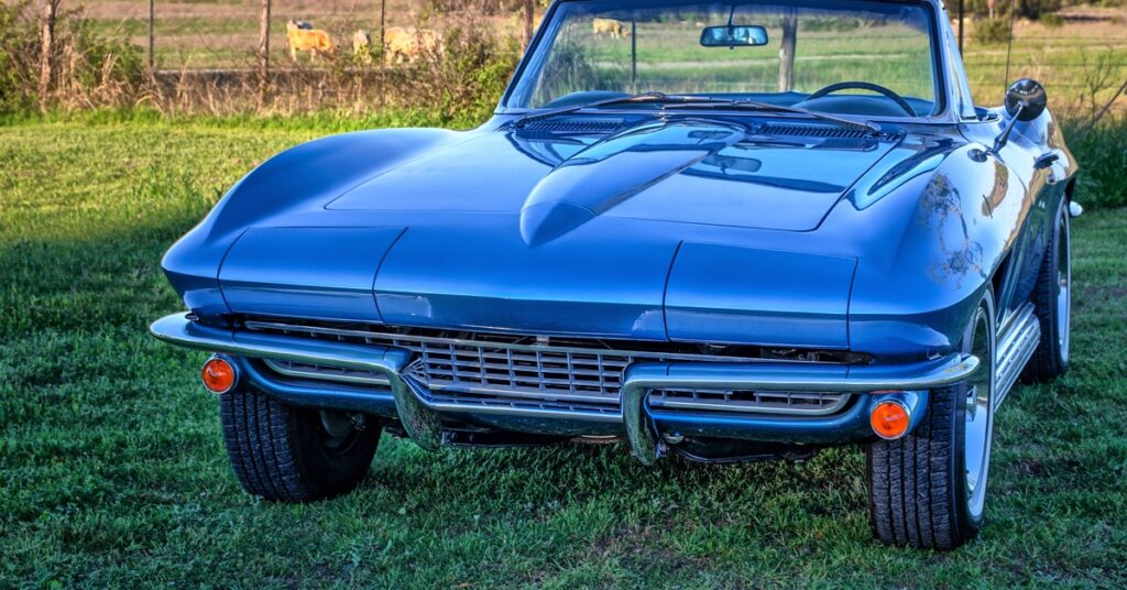Vintage blue convertible car facing camera on grassy field, detailed grille and headlights with countryside backdrop.