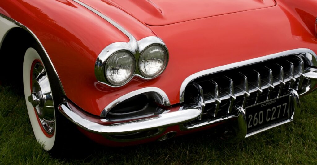 Close-up of red vintage car front with chrome grille, dual headlights, and curved hood parked on grass.