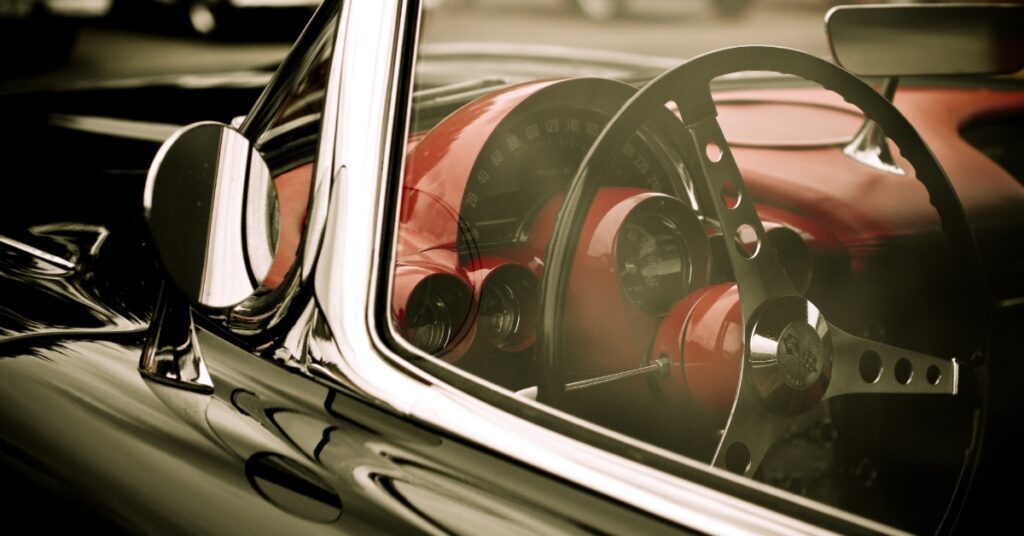 Close-up of classic car interior with red dashboard, circular gauges, and steering wheel seen through side window.