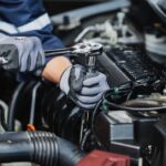 Mechanic in gloves tightens bolt with ratchet inside car engine bay, surrounded by hoses and engine components.
