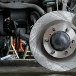 Close-up of a car brake rotor, caliper, and suspension parts with the wheel removed in an auto workshop.