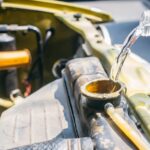 Water flows from a plastic bottle into an open car radiator, with engine hoses and yellow bodywork visible.