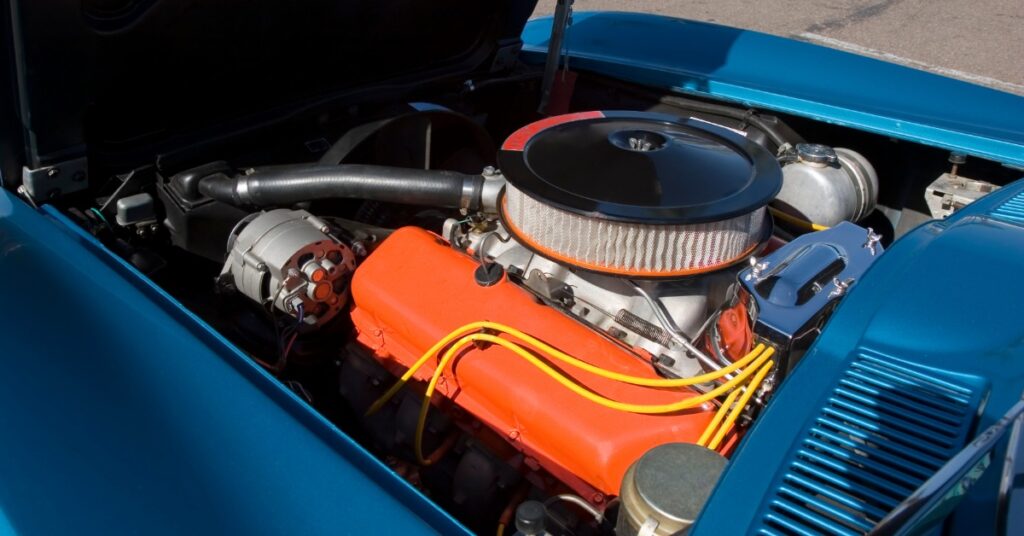 Blue car with its hood raised, revealing an orange engine block, circular air cleaner, yellow wires, and an alternator.