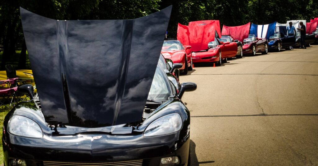 Front view of a black sports car with an open hood, leading a line of red and blue cars with raised hoods outdoors.