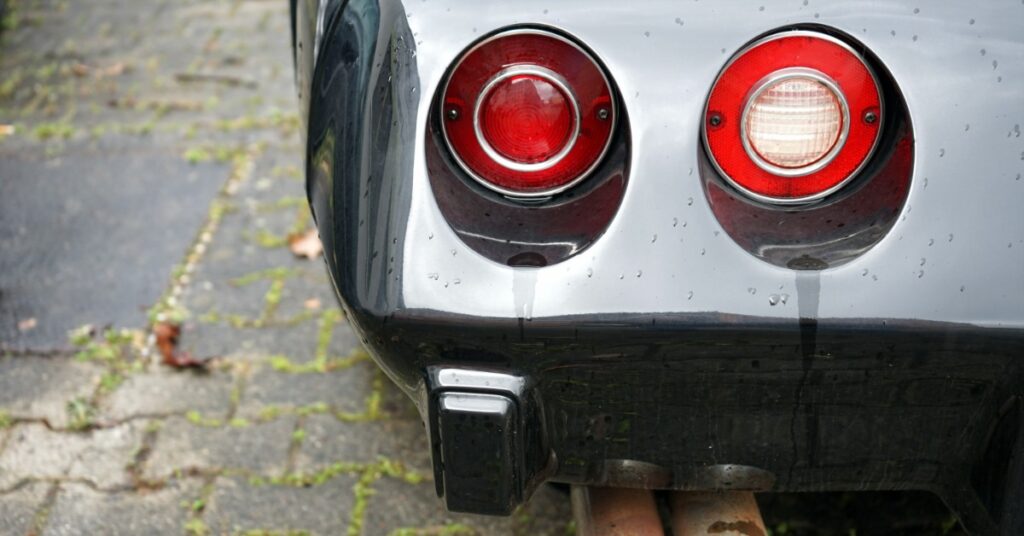 Close-up of twin exhaust pipes beneath a silver vintage car with circular taillights after rain on damp pavement.