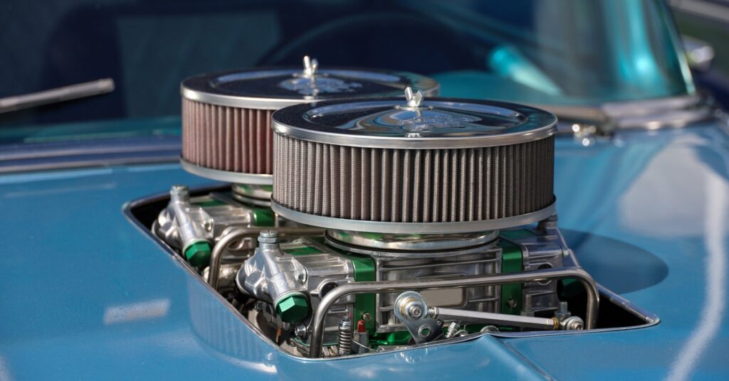 Close-up of a blue classic car with twin round air filters and polished engine hardware visible through the hood.