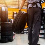 A male auto mechanic is holding two tires in a tire shop's storage room, at a large warehouse within a service center.