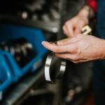 Close-up of a technician in blue overalls measures a metal ring with a caliper beside industrial machine parts.