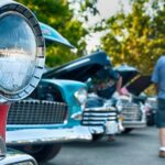 Close-up of a red classic car headlight with other vintage cars and visitors at an outdoor car show.