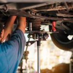 Mechanic works under raised car on lift, reaching toward suspension and wheel components in auto repair shop.