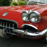 Close view of red vintage convertible with chrome grille and round headlights parked on tree-lined city street.