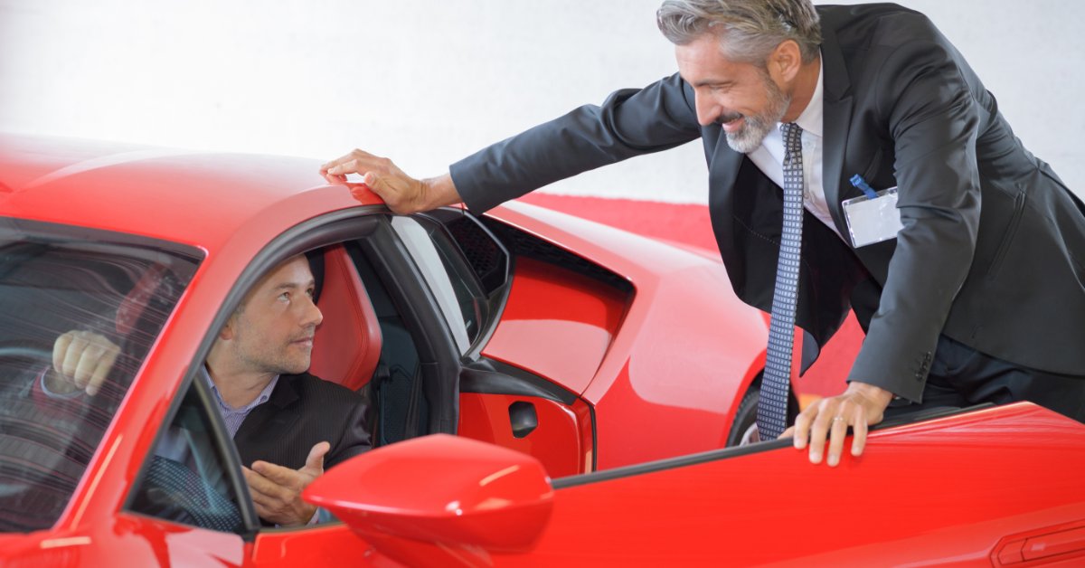 Man in a suit leans into an open red sports car door while speaking with a seated driver in a showroom.
