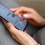 Close-up of hands holding and tapping a smartphone with a blurred app screen while seated on a gray sofa