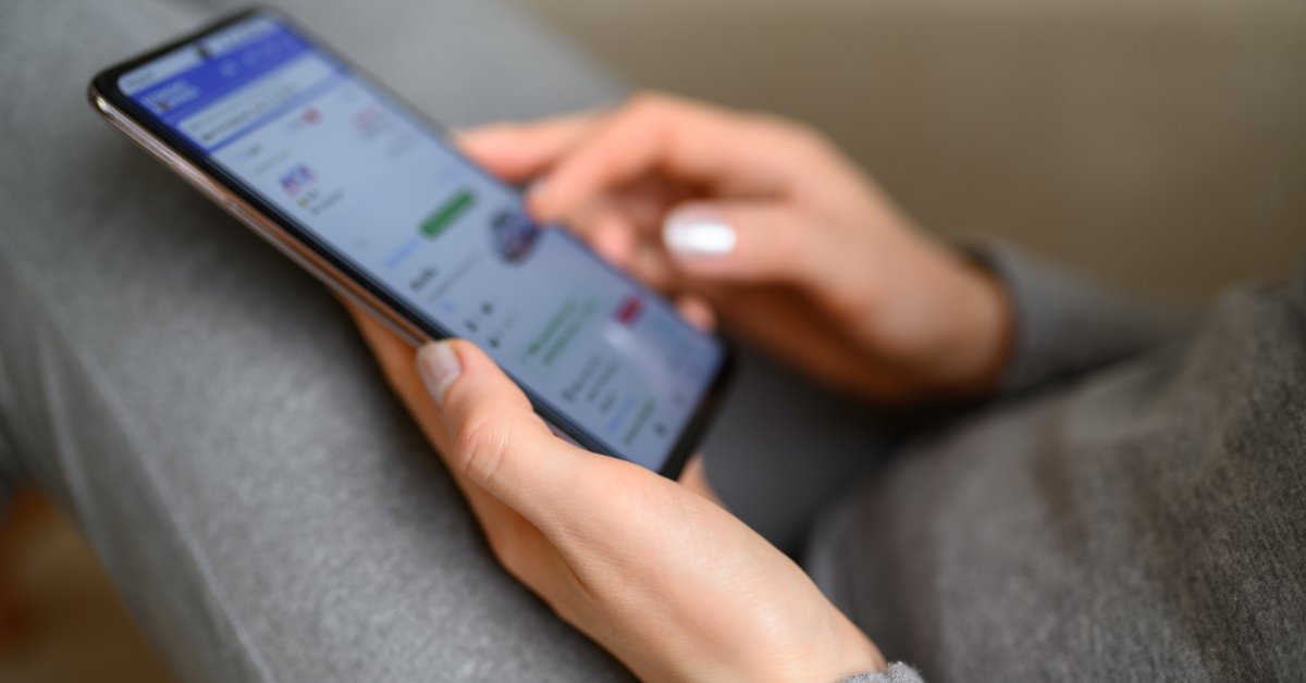 Close-up of hands holding and tapping a smartphone with a blurred app screen while seated on a gray sofa