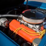 Blue car with its hood raised, revealing an orange engine block, circular air cleaner, yellow wires, and an alternator.