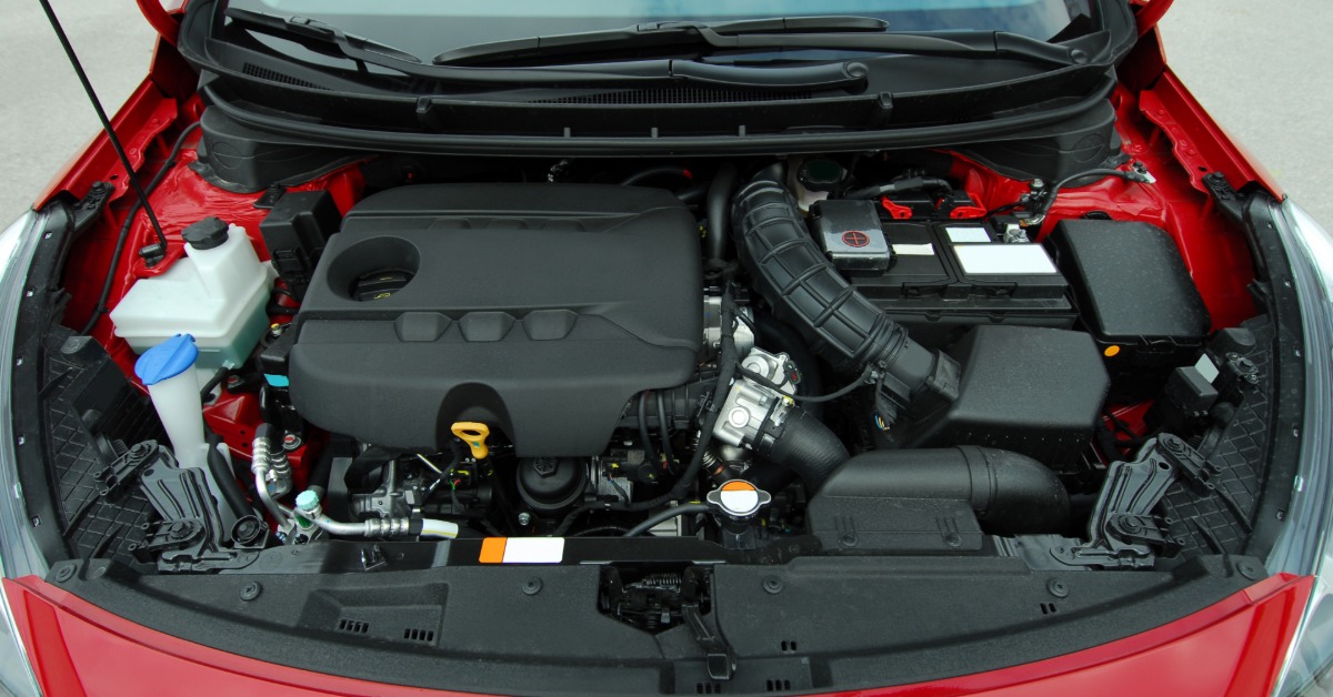 Top view of a red car's engine compartment showing the engine cover, battery, hoses, air intake, and fluid reservoirs.