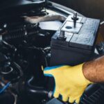 Gloved hands hold a black car battery over an open engine compartment during an automotive repair job.