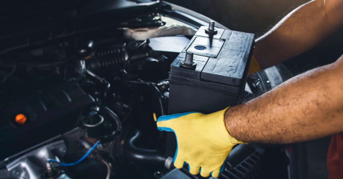 Gloved hands hold a black car battery over an open engine compartment during an automotive repair job.