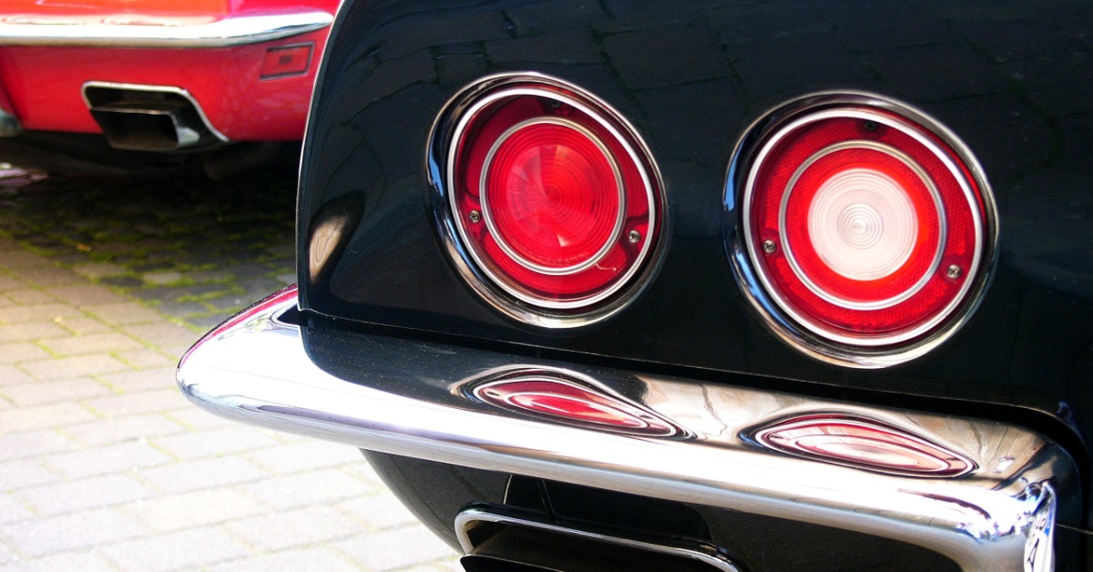 Twin circular taillights and a chrome bumper fill the rear of a black vintage car, with a red car behind it.