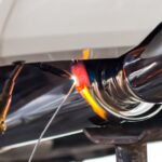 Close-up of a chrome tailpipe being welded under a car as a handheld torch heats the metal bright orange.