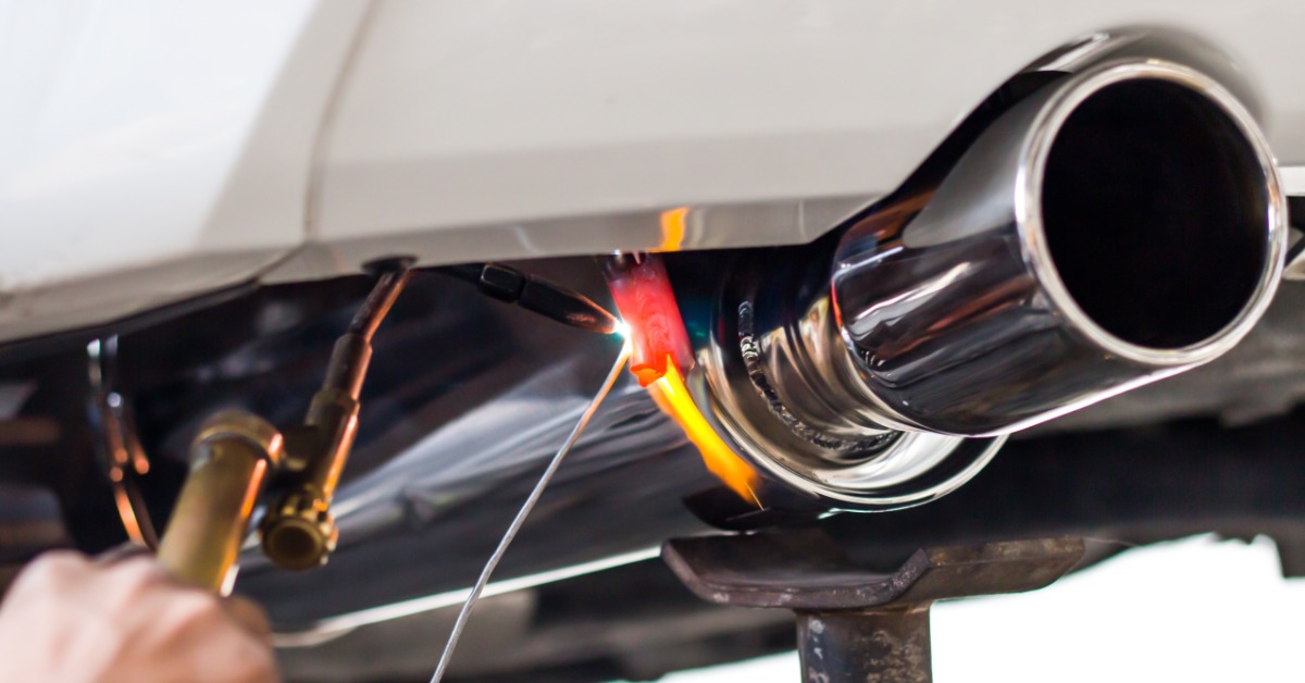 Close-up of a chrome tailpipe being welded under a car as a handheld torch heats the metal bright orange.