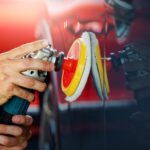 Close-up of hands operating a power polisher with a foam buffing pad on a black car door panel