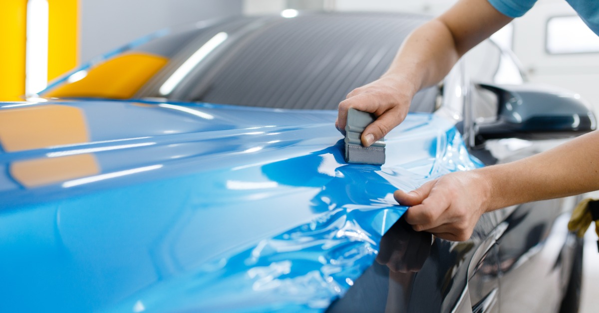 Hands use a squeegee to apply blue vinyl wrap smoothly across the hood of a car inside an automotive detailing shop