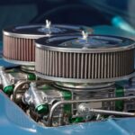 Close-up of a blue classic car with twin round air filters and polished engine hardware visible through the hood.