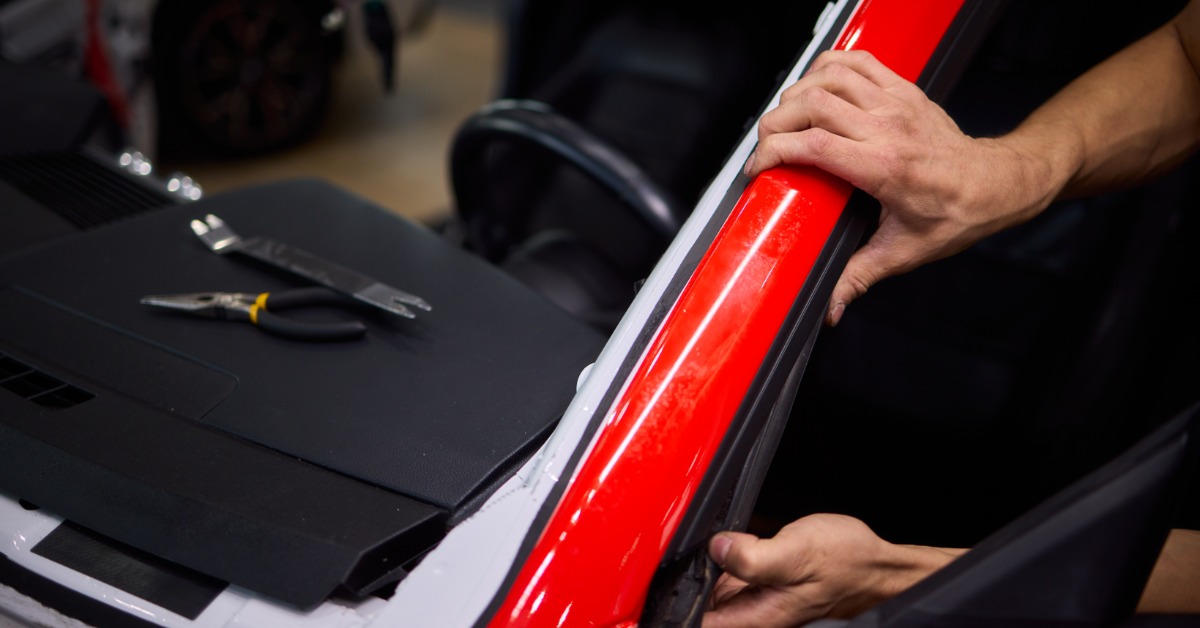 A technician presses black rubber trim along the red frame of a vehicle windshield during repair work.