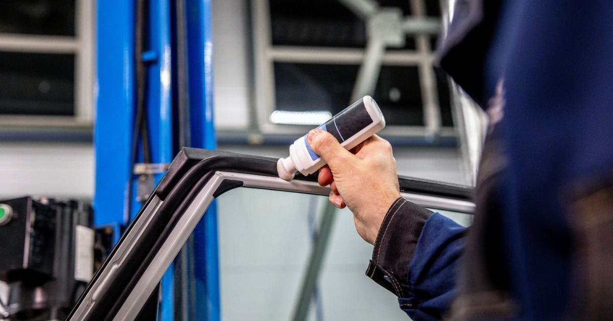 Close-up of a technician applying adhesive to the rubber trim around a vehicle window inside an auto repair shop.