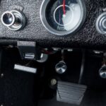 Detailed view of a classic car's black textured dashboard showing labeled switches, a battery meter, and foot pedals.