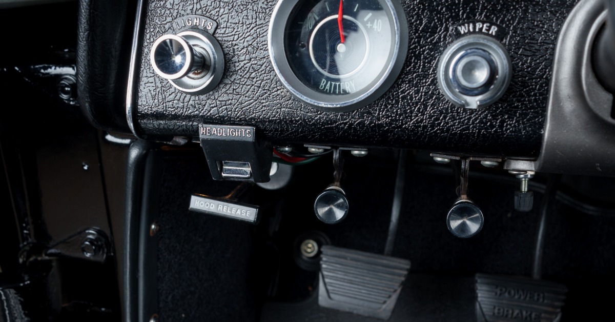 Detailed view of a classic car's black textured dashboard showing labeled switches, a battery meter, and foot pedals.